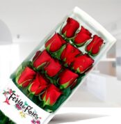 Caja rectangular transparente con 12 rosas rojas con tallos y hojas verdes sobre fondo interior desenfocado