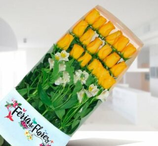 Caja rectangular con 18 rosas amarillas alineadas y flores blancas sobre fondo desenfocado