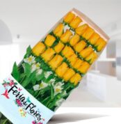 Caja de 24 rosas amarillas ordenadas en filas con follaje verde y flores blancas, envueltas en papel con el logo Feria de Flores