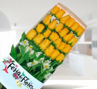 Caja de 24 rosas amarillas ordenadas en filas con follaje verde y flores blancas, envueltas en papel con el logo Feria de Flores