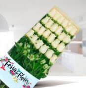 Caja de 24 rosas blancas con follaje verde y envoltura de Feria de Flores sobre fondo interior desenfocado