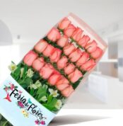 Caja transparente con 24 rosas rosadas en filas ordenadas y follaje verde, con etiqueta Feria de Flores