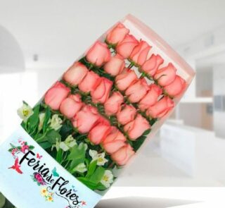 Caja transparente con 24 rosas rosadas en filas ordenadas y follaje verde, con etiqueta Feria de Flores