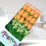 Caja transparente con 12 rosas color salmón y follaje verde de floristería Feria de Flores sobre fondo interior desenfocado