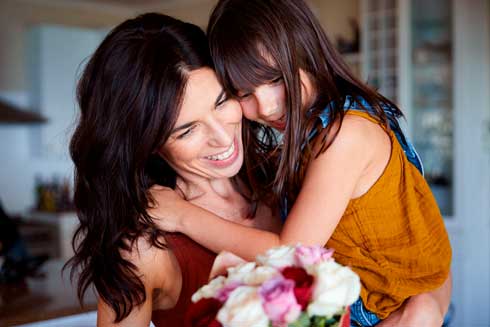 Madre e hija sonrientes se abrazan en casa mientras sostienen un ramo de flores de colores