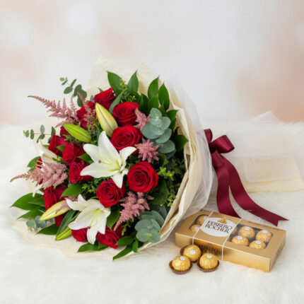 Ramo de rosas rojas con lirios blancos y follaje verde envuelto en papel marfil junto a caja dorada de chocolates Ferrero Rocher sobre superficie blanca