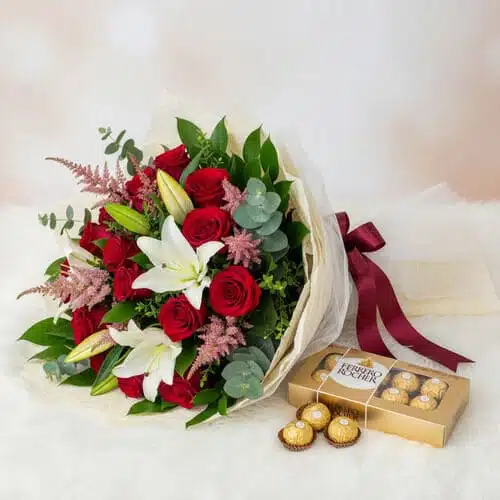 Ramo de rosas rojas con lirios blancos y follaje verde envuelto en papel marfil junto a caja dorada de chocolates Ferrero Rocher sobre superficie blanca