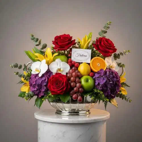 Arreglo de frutas y flores con rosas rojas, hortensias moradas, orquídeas blancas, lirios amarillos y frutas variadas en frutero de vidrio sobre pedestal blanco