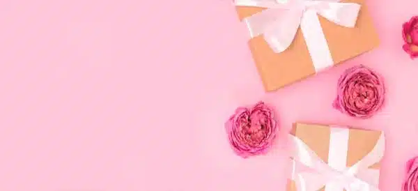 Cajas de regalo color kraft con lazo blanco junto a rosas rosadas sobre fondo rosa pastel