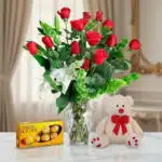 Ramo de rosas rojas en florero de cristal junto a caja de chocolates Ferrero Rocher y oso de peluche blanco con moño rojo sobre mesa