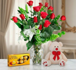 Ramo de rosas rojas en florero de cristal junto a caja de chocolates Ferrero Rocher y oso de peluche blanco con moño rojo sobre mesa