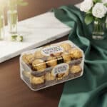 Caja transparente de bombones Ferrero Rocher envueltos en papel dorado sobre mesa de madera con mantel verde y flores blancas al fondo