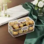 Caja transparente de bombones Ferrero Rocher envueltos en papel dorado sobre mesa de madera con mantel verde y flores blancas al fondo