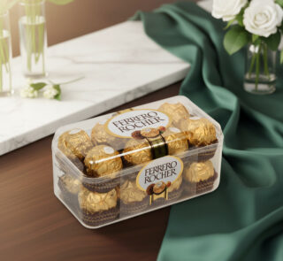 Caja transparente de bombones Ferrero Rocher envueltos en papel dorado sobre mesa de madera con mantel verde y flores blancas al fondo