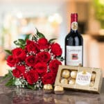 Arreglo de rosas rojas con follaje verde junto a una botella de vino tinto Gato Negro Cabernet Sauvignon y una caja de chocolates Ferrero Rocher sobre una mesa