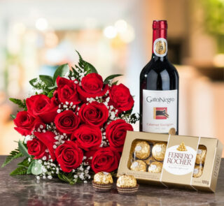 Arreglo de rosas rojas con follaje verde junto a una botella de vino tinto Gato Negro Cabernet Sauvignon y una caja de chocolates Ferrero Rocher sobre una mesa