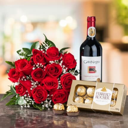 Arreglo de rosas rojas con follaje verde junto a una botella de vino tinto Gato Negro Cabernet Sauvignon y una caja de chocolates Ferrero Rocher sobre una mesa