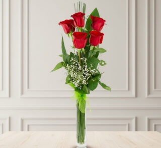 ramo vertical de seis rosas rojas con follaje verde y paniculata en florero cilíndrico de cristal sobre mesa de madera clara