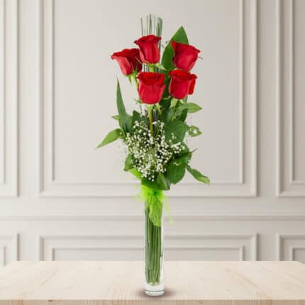 ramo vertical de seis rosas rojas con follaje verde y paniculata en florero cilíndrico de cristal sobre mesa de madera clara
