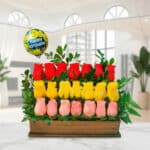 Arreglo floral rectangular con rosas rojas, amarillas y rosadas en base de madera, hojas verdes y globo Happy Birthday en fondo de sala moderna