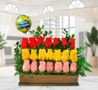 Arreglo floral rectangular con rosas rojas, amarillas y rosadas en base de madera, hojas verdes y globo Happy Birthday en fondo de sala moderna