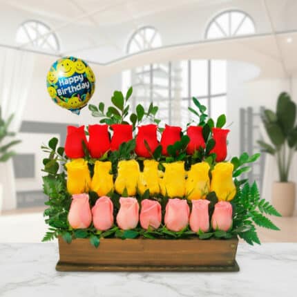 Arreglo floral rectangular con rosas rojas, amarillas y rosadas en base de madera, hojas verdes y globo Happy Birthday en fondo de sala moderna