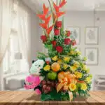 Arreglo de flores amarillas y rojas con heliconias, peluche de vaca blanco y negro, frutas frescas y lazo naranja sobre mesa de madera en sala luminosa