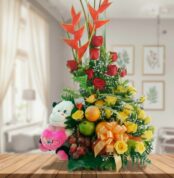 Arreglo de flores amarillas y rojas con heliconias, peluche de vaca blanco y negro, frutas frescas y lazo naranja sobre mesa de madera en sala luminosa