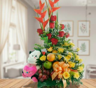 Arreglo de flores amarillas y rojas con heliconias, peluche de vaca blanco y negro, frutas frescas y lazo naranja sobre mesa de madera en sala luminosa