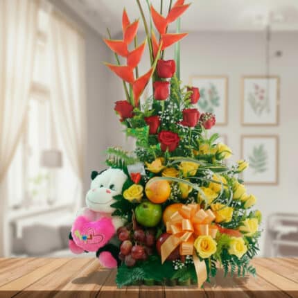 Arreglo de flores amarillas y rojas con heliconias, peluche de vaca blanco y negro, frutas frescas y lazo naranja sobre mesa de madera en sala luminosa