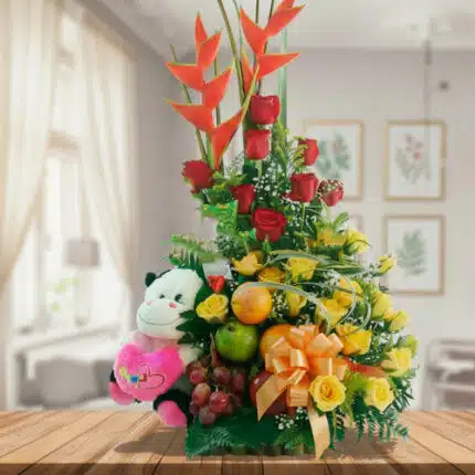 Arreglo de flores amarillas y rojas con heliconias, peluche de vaca blanco y negro, frutas frescas y lazo naranja sobre mesa de madera en sala luminosa
