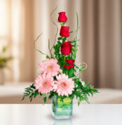 Arreglo vertical de rosas rojas y gerberas rosadas en un florero cuadrado de vidrio sobre una mesa blanca, con fondo desenfocado en tonos beige