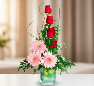 Arreglo vertical de rosas rojas y gerberas rosadas en un florero cuadrado de vidrio sobre una mesa blanca, con fondo desenfocado en tonos beige