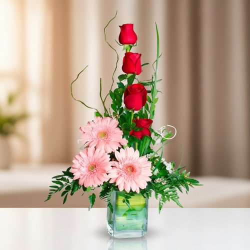 Arreglo floral con rosas rojas y gerberas rosadas en florero de vidrio Arreglo vertical de rosas rojas y gerberas rosadas en un florero cuadrado de vidrio sobre una mesa blanca, con fondo desenfocado en tonos beige