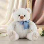 Osito de peluche blanco y suave con lazo azul sentado sobre una superficie clara, con cortinas y marco dorado de fondo