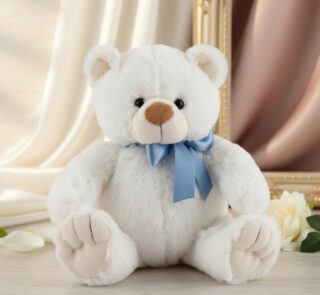 Osito de peluche blanco y suave con lazo azul sentado sobre una superficie clara, con cortinas y marco dorado de fondo
