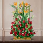 arreglo floral rectangular con rosas rojas, lirios blancos y aves del paraíso naranjas sobre base de madera frente a pared clásica