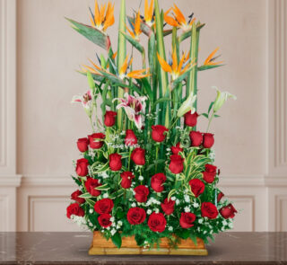 arreglo floral rectangular con rosas rojas, lirios blancos y aves del paraíso naranjas sobre base de madera frente a pared clásica