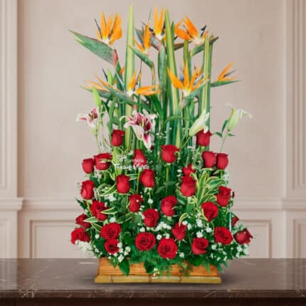 arreglo floral rectangular con rosas rojas, lirios blancos y aves del paraíso naranjas sobre base de madera frente a pared clásica