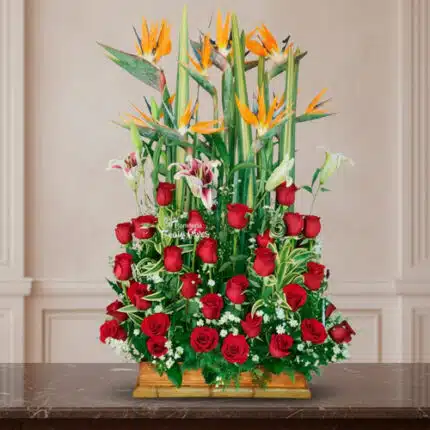 arreglo floral rectangular con rosas rojas, lirios blancos y aves del paraíso naranjas sobre base de madera frente a pared clásica