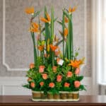 Arreglo floral alto con aves del paraíso y rosas naranjas en base de bambú sobre mesa interior