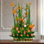 Arreglo floral alto con aves del paraíso y rosas naranjas en base de bambú sobre mesa interior