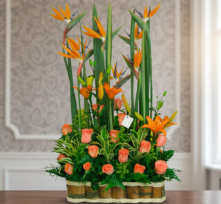 Arreglo floral alto con aves del paraíso y rosas naranjas en base de bambú sobre mesa interior