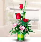 arreglo floral vertical con tres rosas rojas, gerberas rosas y follaje verde en jarrón de vidrio cilíndrico sobre fondo de sala desenfocado