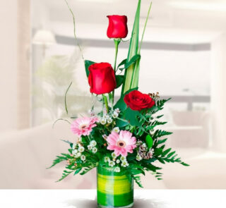 arreglo floral vertical con tres rosas rojas, gerberas rosas y follaje verde en jarrón de vidrio cilíndrico sobre fondo de sala desenfocado