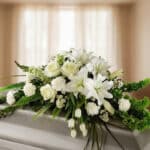 Centro de flores blancas con lirios, rosas y claveles sobre un ataúd en una sala iluminada