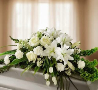 Centro de flores blancas con lirios, rosas y claveles sobre un ataúd en una sala iluminada