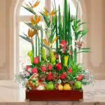 Arreglo de flores tropicales y rosas rojas combinado con frutas variadas en una caja de madera sobre una mesa de mármol, con fondo interior desenfocado