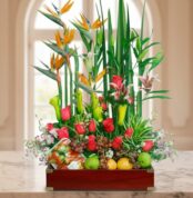 Arreglo de flores tropicales y rosas rojas combinado con frutas variadas en una caja de madera sobre una mesa de mármol, con fondo interior desenfocado