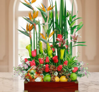 Arreglo de flores tropicales y rosas rojas combinado con frutas variadas en una caja de madera sobre una mesa de mármol, con fondo interior desenfocado